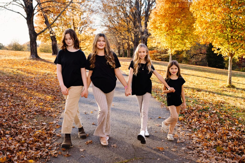 Family photographer, sisters holding hands walking in line on fall trail