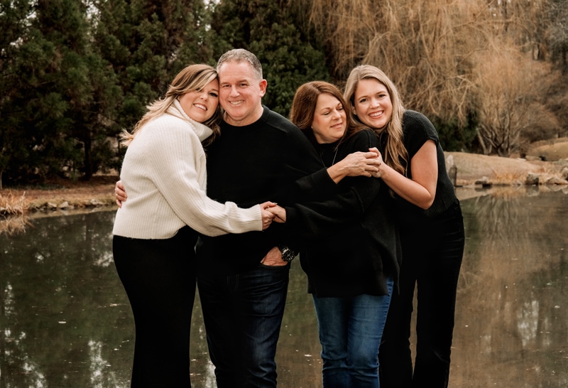 Family photographer, mom and dad hugging daughters on bridge over pond
